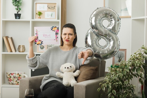 Unzufrieden mit Daumen nach unten schönes Mädchen am glücklichen Frauentag mit Grußkarte auf Sessel im Wohnzimmer sitzend