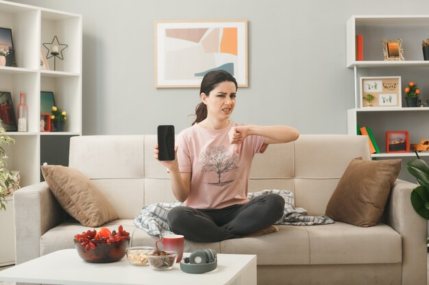 Unzufrieden mit dem Daumen nach unten junges Mädchen, das Telefon hält, das auf dem Sofa hinter dem Couchtisch im Wohnzimmer sitzt