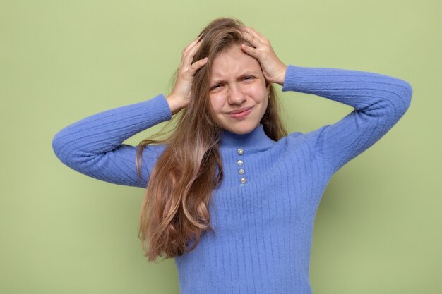 Unzufrieden packte den Kopf schönes kleines Mädchen mit blauem Pullover isoliert auf olivgrüner Wand
