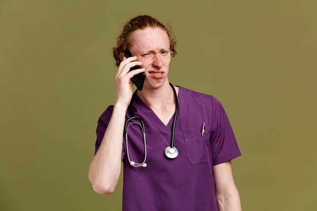 Unzufrieden spricht am Telefon junger männlicher Arzt in Uniform mit Stethoskop isoliert auf grünem Hintergrund