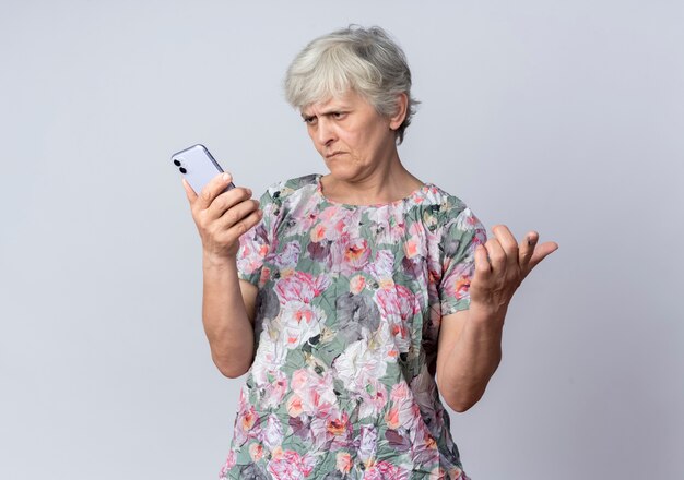Unzufriedene ältere Frau hält und betrachtet Telefon isoliert auf weißer Wand