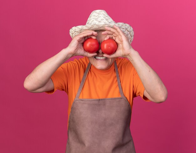 Unzufriedene ältere Gärtnerin mit Gartenhut, die die Augen mit Tomaten bedeckt