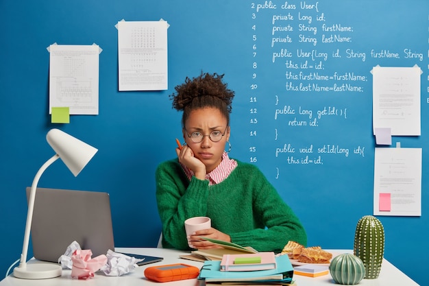 Unzufriedene Afro-Frau hält Stift, hat die Prüfungsvorbereitung satt, trinkt Kaffee und schaut traurig in die Kamera, sitzt mit der notwendigen Ausrüstung und dem Notizblock am Schreibtisch zu Hause