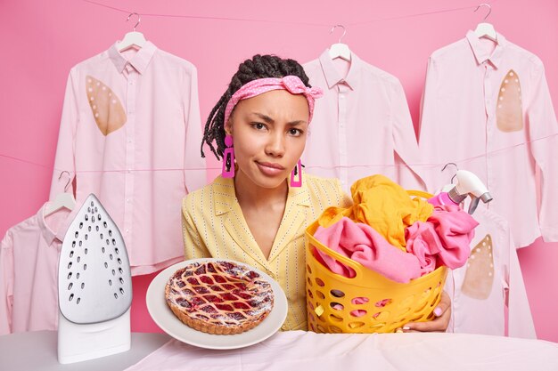 Unzufriedene afroamerikanische Hausfrau, die zu Hause mit Kochen, Waschen und Bügeln beschäftigt ist, macht Haushaltsaufgaben gegen gebügelte Kleidung, die an Seilständern in der Nähe des Brettes hängt.