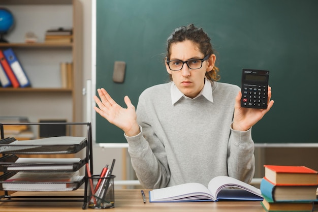 unzufriedene Ausbreitung der Hände junger männlicher Lehrer, der einen Taschenrechner hält, der am Schreibtisch mit Schulwerkzeugen im Klassenzimmer sitzt