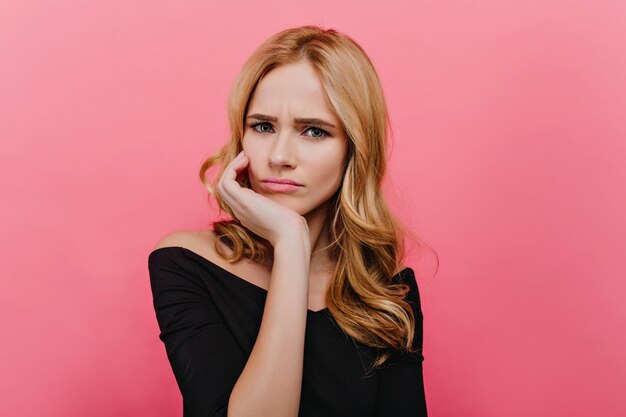 Unzufriedene blauäugige Dame, die lustige Gesichter auf heller Wand macht. Faszinierende lockige Frau in schwarzer Kleidung, die Traurigkeit aufwirft und ausdrückt.