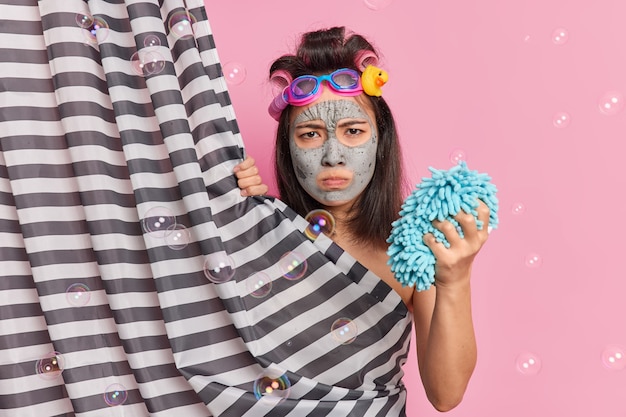 Unzufriedene düstere asiatische Frau mit dunklem Haar macht Frisur angewendet Gesichtsmaske für die Hautverjüngung hält Duschschwamm hat wütende Stimmung posiert hinter Vorhang isoliert auf rosa Studiowand