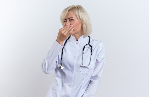 Unzufriedene erwachsene ärztin in medizinischer robe mit stethoskop, die ihre nase isoliert auf weißer wand mit kopienraum hält