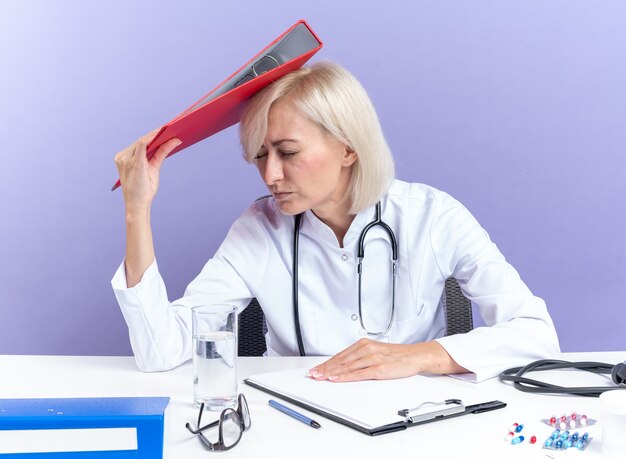Unzufriedene erwachsene slawische Ärztin in medizinischer Robe mit Stethoskop am Schreibtisch sitzend mit Bürowerkzeugen, die Dateiordner auf dem Kopf halten, isoliert auf violettem Hintergrund mit Kopienraum