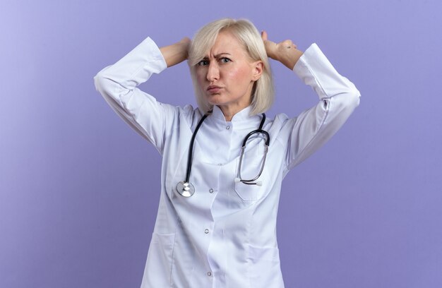 Unzufriedene erwachsene slawische Ärztin in medizinischer Robe mit Stethoskop, die mit erhobenen Händen auf violettem Hintergrund mit Kopienraum isoliert steht
