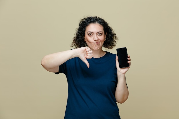 unzufriedene Frau mittleren Alters, die ein T-Shirt trägt, das ein Handy zeigt, das in die Kamera schaut und den Daumen nach unten zeigt, isoliert auf olivgrünem Hintergrund