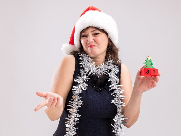 Unzufriedene Frau mittleren Alters mit Weihnachtsmütze und Lametta-Girlande um den Hals, die ein Weihnachtsbaumspielzeug mit Datum hält und auf die Seite zeigt, isoliert auf weißem Hintergrund on