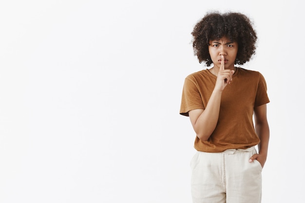 Unzufriedene herrische Afroamerikanerin mit Afro-Frisur, die die Stirn runzelt, weil sie nicht gerne sagt, dass sie eine Shush-Geste mit dem Zeigefinger über dem geschlossenen Mund zeigt