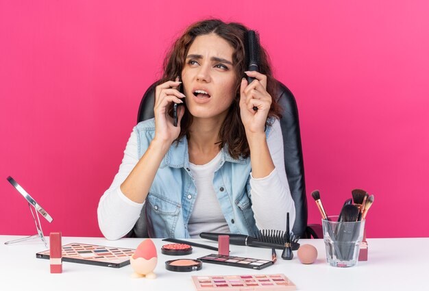 Unzufriedene hübsche kaukasische Frau, die am Tisch mit Make-up-Tools sitzt und am Telefon spricht und ihr Haar kämmt