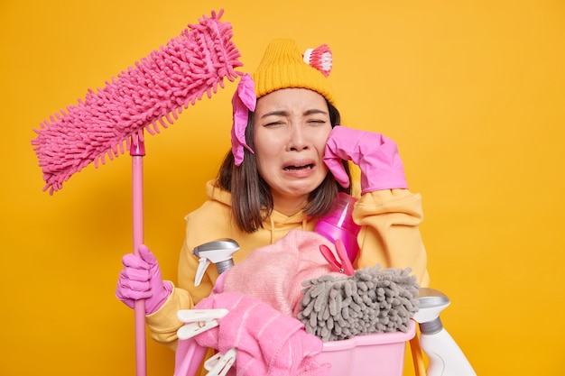 Kostenloses Foto unzufriedene junge asiatin fühlt sich sehr müde weint, da sie nicht mit der hausreinigung fortfahren möchte, hält mop lehnt sich mit schmutziger wäsche an den korb und reinigungsmittel hat eine frist, um das zimmer aufzuräumen?