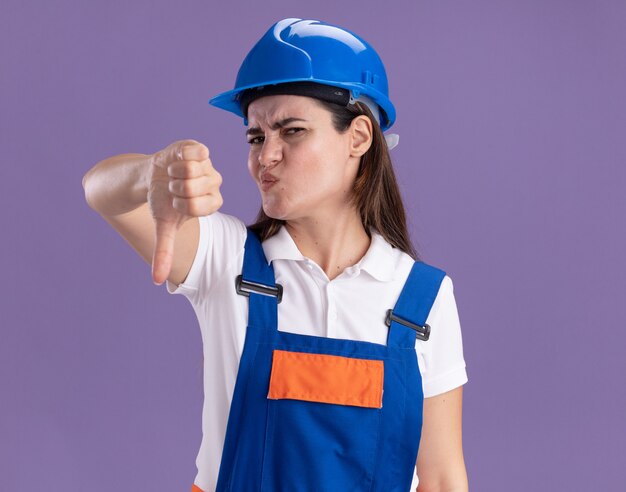 Unzufriedene junge Baumeisterin in Uniform mit Daumen nach unten isoliert auf lila Wand