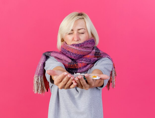Unzufriedene junge blonde kranke slawische Frau, die Schal trägt, hält Packungen mit medizinischen Pillen
