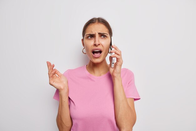 Unzufriedene junge brünette Frau hat einen verärgerten Ausdruck, runzelt die Stirn, ruft vor Ärger aus, hat ein langweiliges Telefongespräch, einen verwirrten Blick und trägt ein lässiges rosa T-Shirt, das über weißem Hintergrund isoliert ist