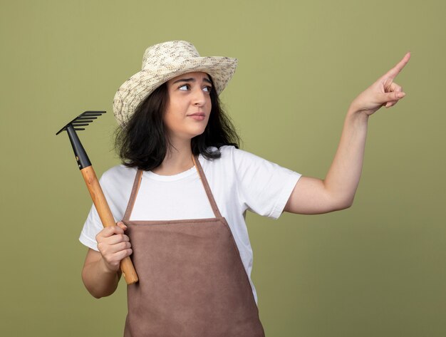 Unzufriedene junge brünette weibliche Gärtnerin in Uniform, die Gartenhut trägt, hält Rechen, der auf Seite lokalisiert auf olivgrüne Wand schaut und zeigt