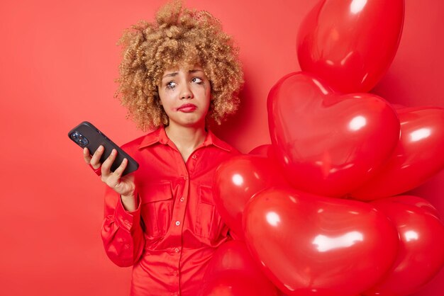 Unzufriedene junge Europäerin hat die Stimmung verdorben, wartet auf den Valentinstag, benutzt das Handy, fühlt sich einsam, hält einen Haufen Herzballons, hat Make-up isoliert über rotem Hintergrund durchgesickert