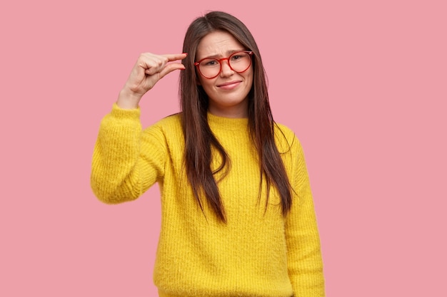 Unzufriedene junge Frau hat langes Haar, beschreibt Größe mit den Fingern, zeigt etwas Kleines, trägt eine optische Brille, gelben warmen Pullover