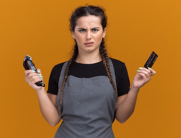 Unzufriedene junge Friseurin in Uniform mit Kreditkarte und Haarschneidemaschine isoliert auf oranger Wand