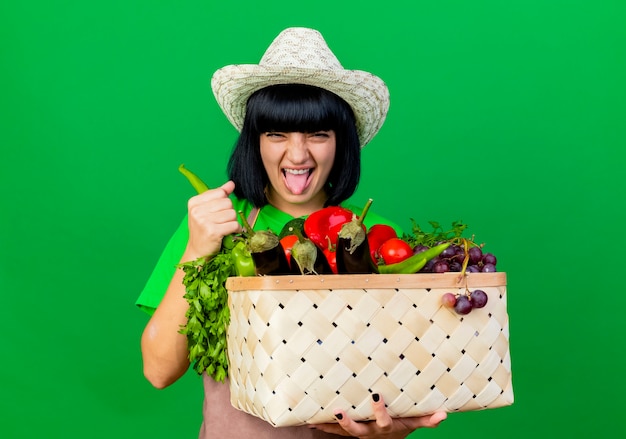 Unzufriedene junge Gärtnerin in Uniform mit Gartenhut hält Gemüsekorb und Paprika