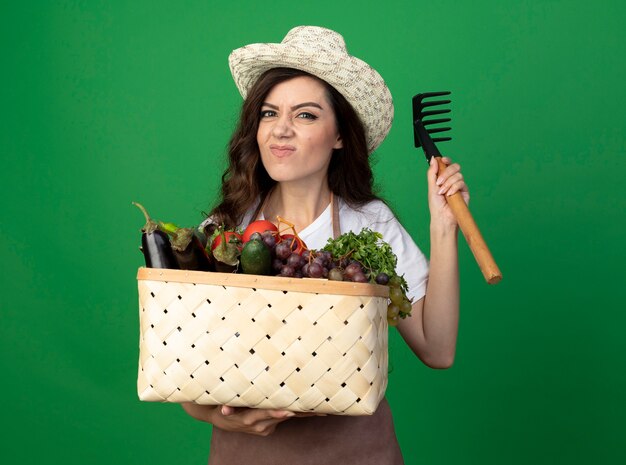 Unzufriedene junge Gärtnerin in Uniform mit Gartenhut hält Gemüsekorb und Rechen isoliert auf grüner Wand