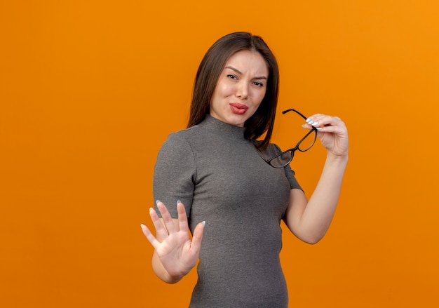 Unzufriedene junge hübsche Frau, die Brille hält und nicht isoliert auf orange Hintergrund mit Kopienraum gestikuliert
