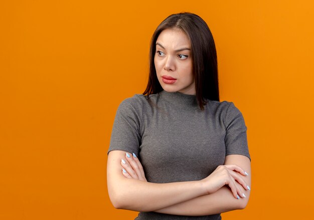 Unzufriedene junge hübsche Frau, die Seite betrachtet und mit geschlossener Haltung steht, lokalisiert auf orange Hintergrund mit Kopienraum