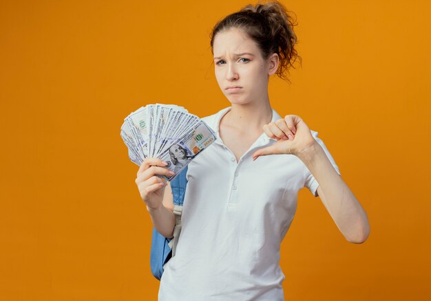 Unzufriedene junge hübsche Studentin, die Rückentasche hält, die Geld hält und Daumen unten auf orange Hintergrund mit Kopienraum zeigt