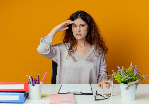 Unzufriedene junge hübsche weibliche Büroangestellte, die am Schreibtisch mit Bürowerkzeugen sitzt, die Hand auf Stirn lokalisiert auf Orange setzen