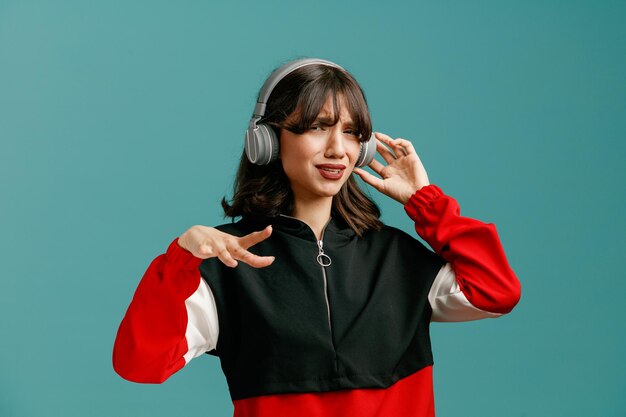 Unzufriedene junge kaukasische Frau, die Kopfhörer trägt und ergreift und die Hand in der Luft hält und isoliert auf blauem Hintergrund in die Kamera blickt
