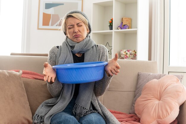 Unzufriedene junge kranke slawische Frau mit Schal um den Hals mit Wintermütze, die einen Eimer hält, der mit geschlossenen Augen im Wohnzimmer auf der Couch sitzt