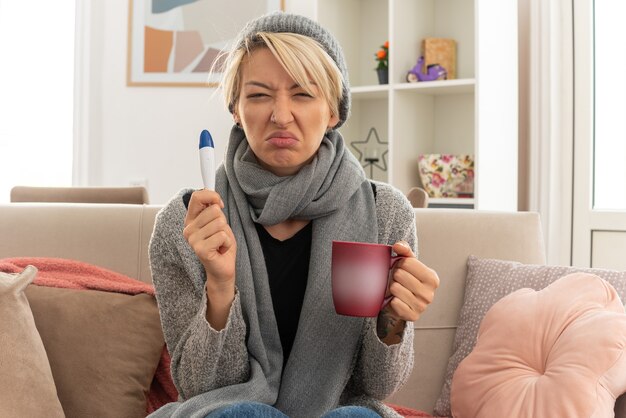 Unzufriedene junge kranke slawische Frau mit Schal um den Hals mit Wintermütze mit Thermometer und Tasse auf der Couch im Wohnzimmer sitzend