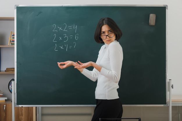 Unzufriedene junge Lehrerin mit Brille, die vor der Tafel steht und im Klassenzimmer schreibt
