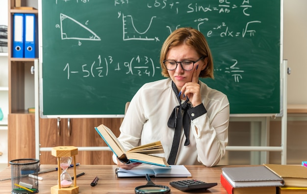 Unzufriedene junge Lehrerin mit Brille sitzt am Tisch mit Schulwerkzeugen, die ein Buch im Klassenzimmer halten und lesen