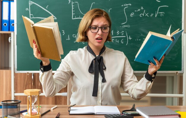 Unzufriedene junge Lehrerin mit Brille sitzt am Tisch mit Schulwerkzeugen, die ein Buch im Klassenzimmer lesen