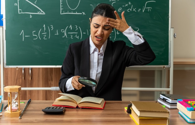 Unzufriedene junge lehrerin sitzt am tisch mit schulmaterial und liest buch mit lupe, die hand auf die stirn im klassenzimmer legt