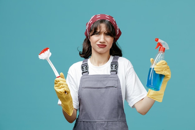 Unzufriedene junge Putzfrau mit einheitlichem Bandana und Gummihandschuhen, die Bürste und Reinigungsmittel zeigen, die isoliert auf blauem Hintergrund in die Kamera blicken
