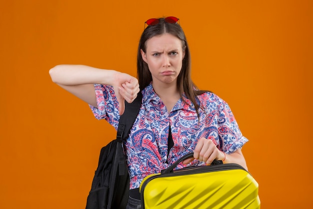 Unzufriedene junge Reisende Frau, die rote Sonnenbrille auf Kopf stehend mit Rucksack hält Koffer mit wütendem Ausdruck zeigt Daumen nach unten