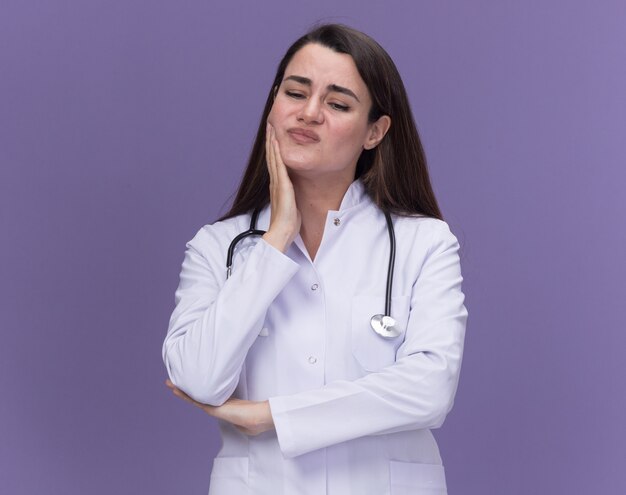 Unzufriedene junge Ärztin, die ein medizinisches Gewand mit Stethoskop trägt, legt die Hand auf das Kinn und schaut isoliert auf lila Wand mit Kopienraum nach unten