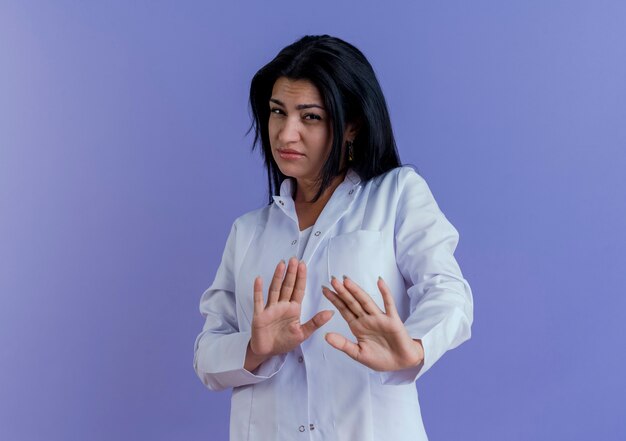 Unzufriedene junge Ärztin, die medizinische Robe trägt, die keine Geste tut, die auf lila Wand mit Kopienraum isoliert wird