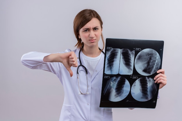 Unzufriedene junge Ärztin, die medizinische Robe und Stethoskop trägt und Röntgenaufnahme hält, die Daumen unten auf isolierter weißer Wand zeigt