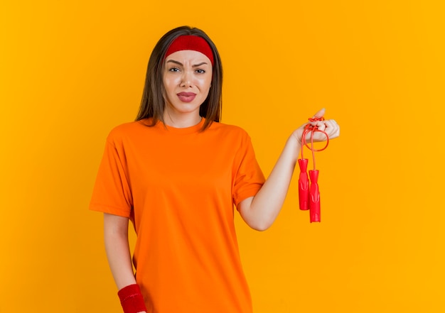 Unzufriedene junge sportliche Frau, die Stirnband und Armbänder hält, die Springseil lokalisiert auf orange Wand mit Kopienraum halten
