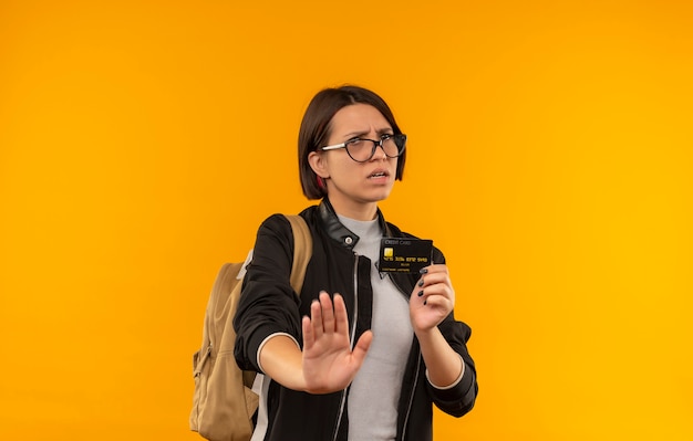 Unzufriedene junge Studentin, die Brille und Rückentasche hält, die Kreditkarten gestikulierenden Halt lokalisiert auf orange Hintergrund mit Kopienraum hält