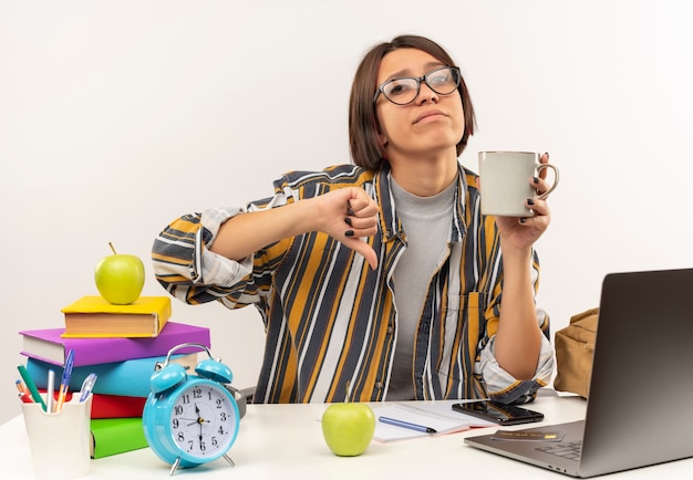 Kostenloses Foto unzufriedene junge studentin, die eine brille trägt, die am schreibtisch mit universitätswerkzeugen sitzt, die tasse kaffee halten und daumen unten lokalisiert auf weißem hintergrund zeigt