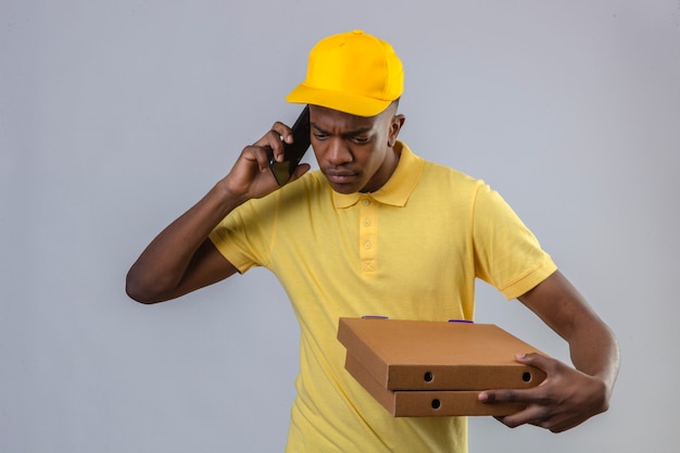 Unzufriedene Lieferung Afroamerikaner Mann in gelbem Poloshirt und Kappe, die Pizzaschachteln hält, während auf Handy sprechen