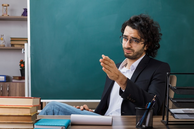 Unzufriedene Punkte an der Seite männlicher Lehrer mit Brille, die am Tisch mit Schulwerkzeugen im Klassenzimmer sitzt