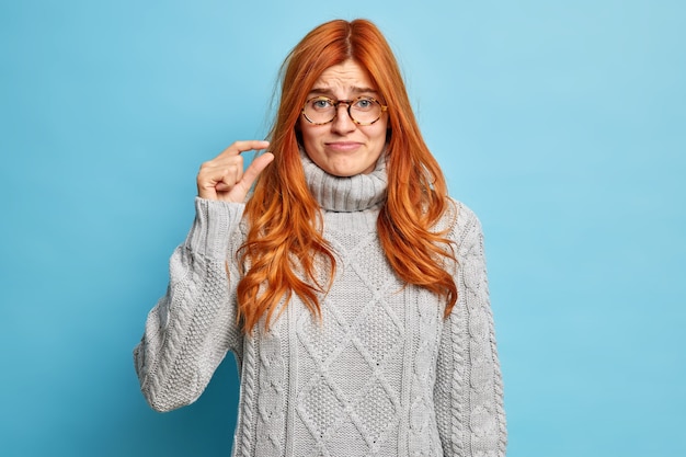 Unzufriedene rothaarige junge europäische Frau in grauem Winterpullover zeigt kleine Menge Geste zeigt winzige Größe mit den Fingern.